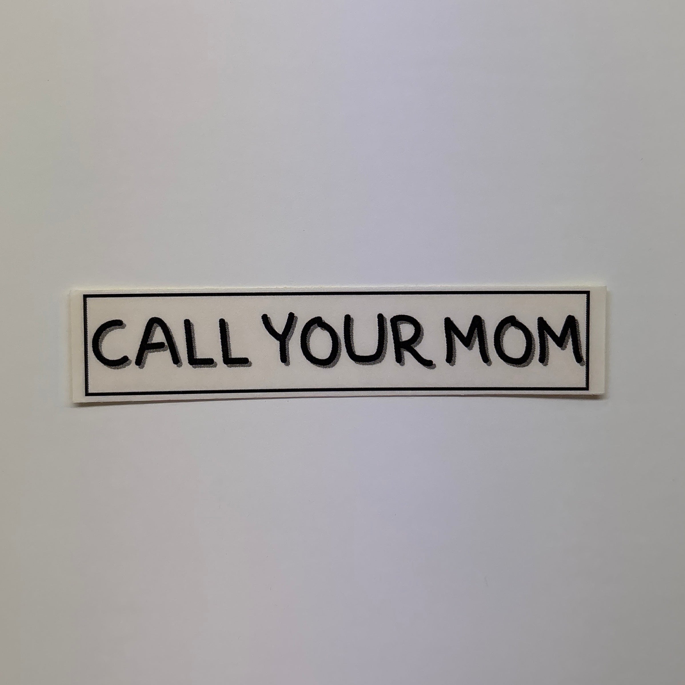 White surface with a black text sticker reading 'CALL YOUR MOM' on a plain background