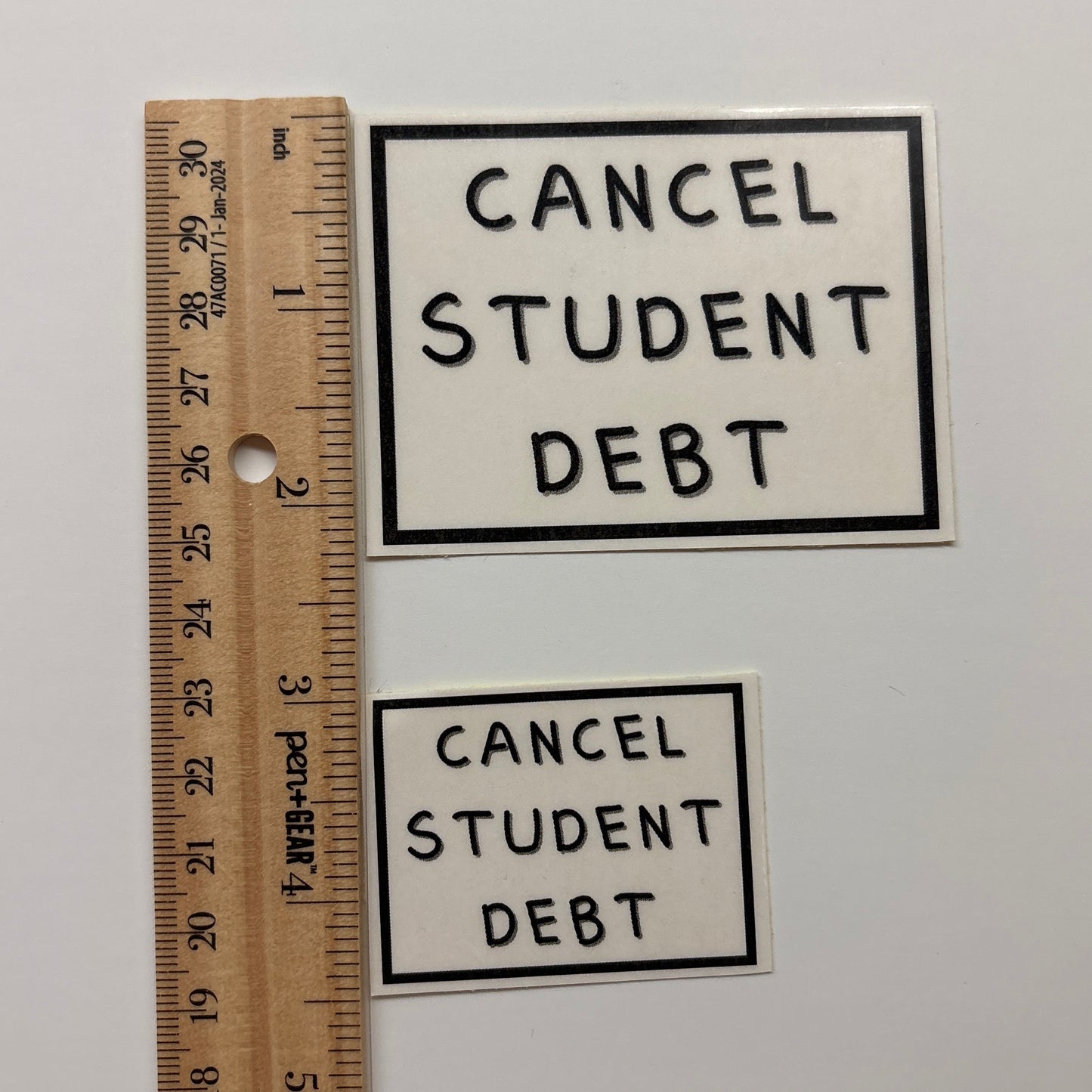 Two 'Cancel Student Debt' stickers with a ruler for scale on a white background