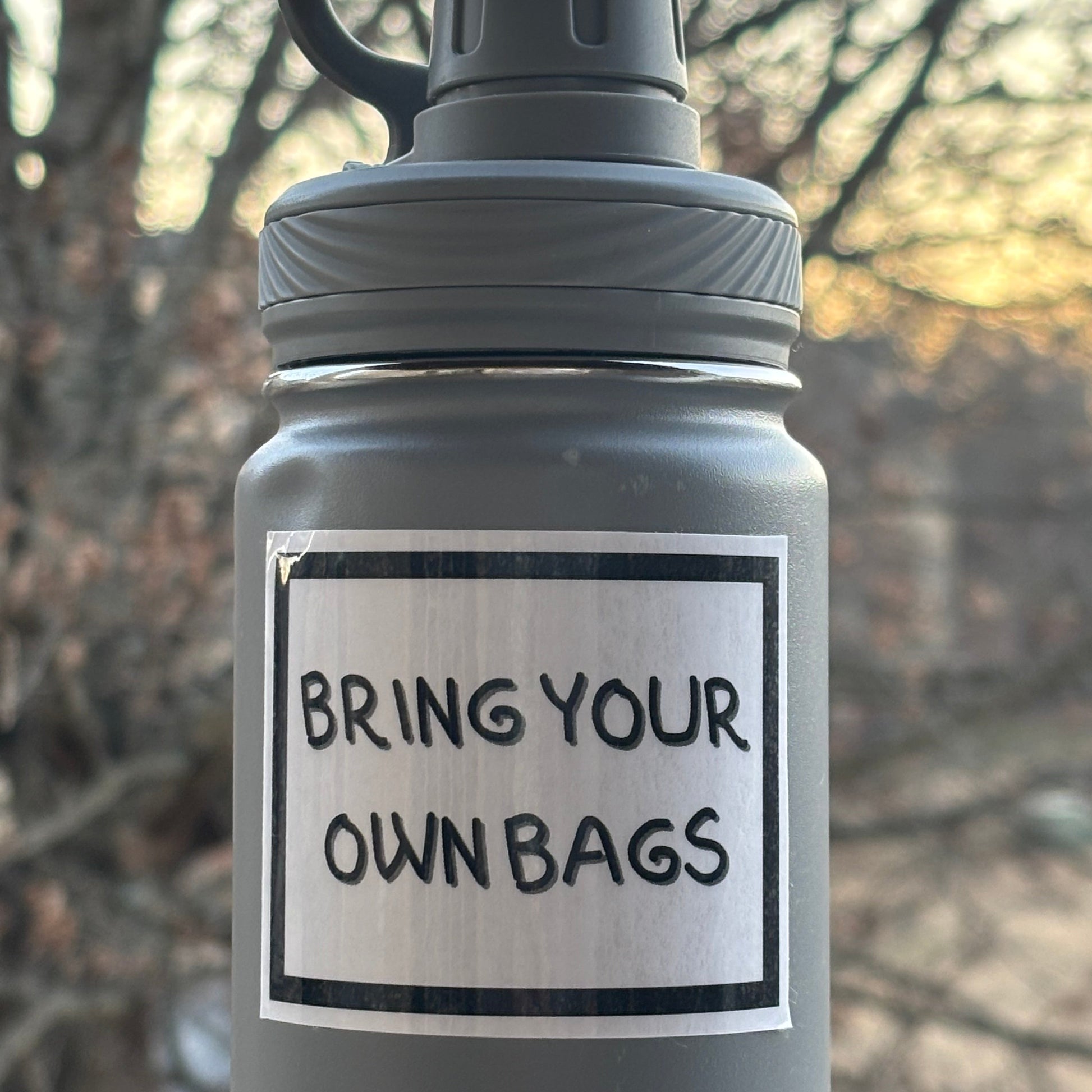 Gray water bottle with 'Bring Your Own Bags' label against a blurred natural background