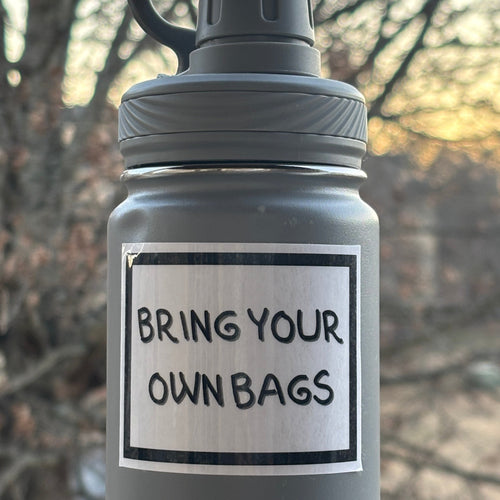 Gray water bottle with 'Bring Your Own Bags' label against a blurred natural background