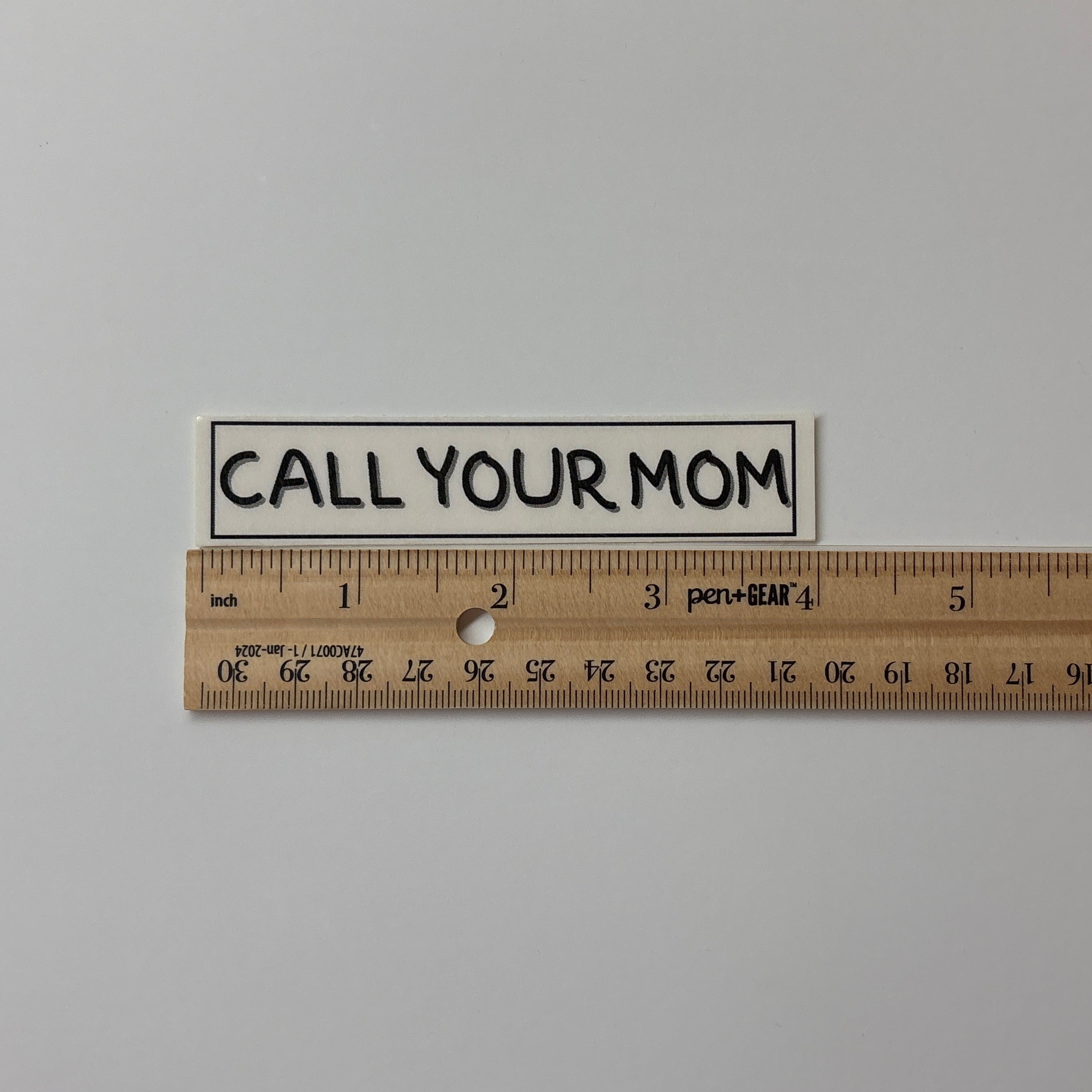 Small sign with 'CALL YOUR MOM' text next to a ruler on a plain background